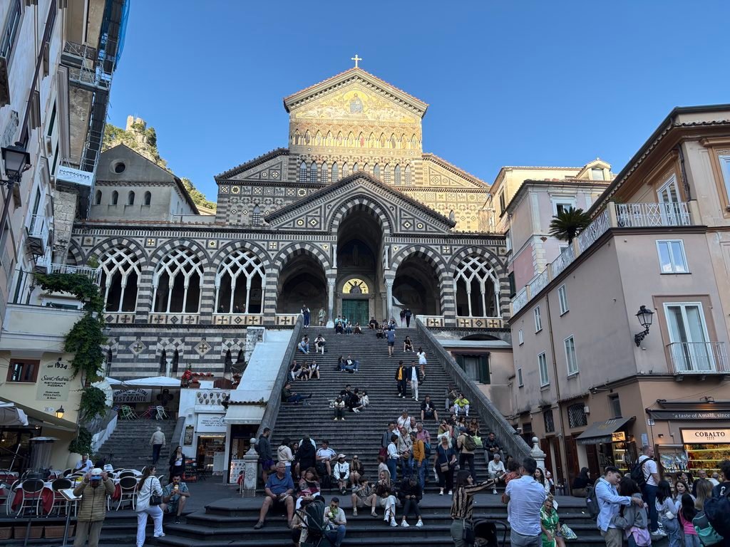 Front of Amalfi Cathedral with its striped arches, broad staircase and people gathered in the square.