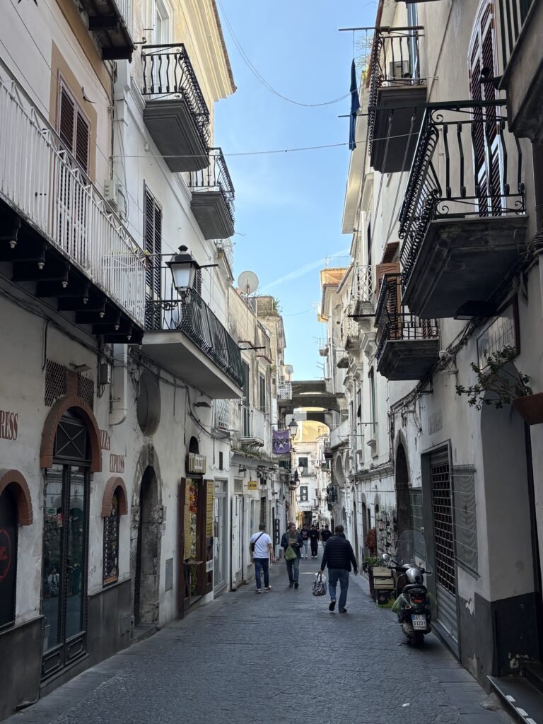 Narrow pedestrian street in Amalfi lined with buildings, balconies and small shops.