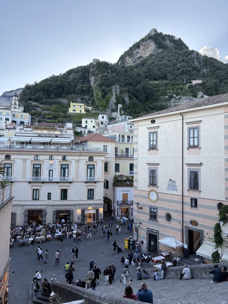 View over Amalfi's main square with restaurants, pedestrians and buildings below the surrounding hillside.