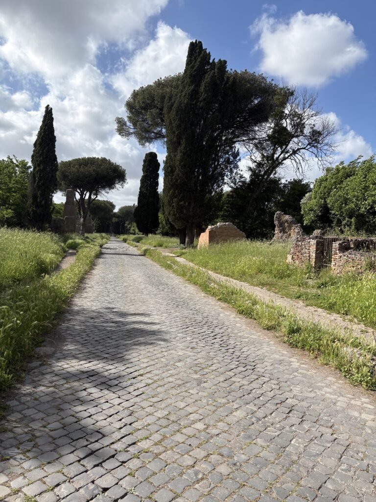 Stone-paved Appian Way lined with grass, cypress trees and ancient ruins.