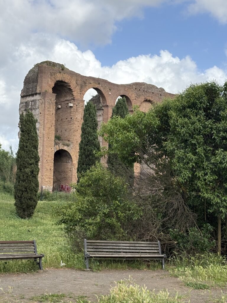 Ancient brick aqueduct ruins beside benches and green parkland in Rome.