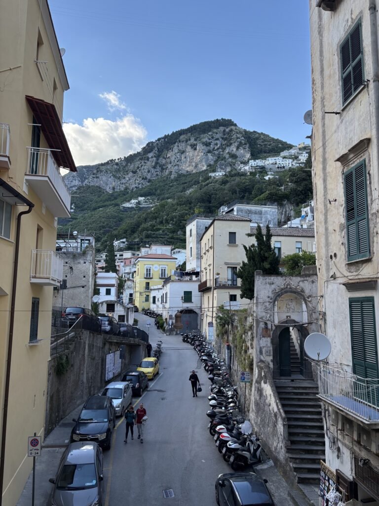 Road through Amalfi with parked cars and scooters, buildings on both sides and mountains behind.