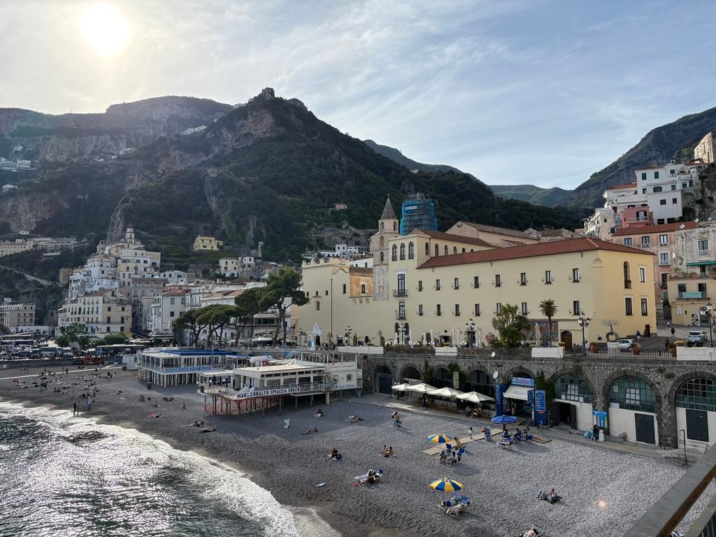 View of Amalfi Town from the waterfront, with the beach, harbour buildings and steep mountains in the background.